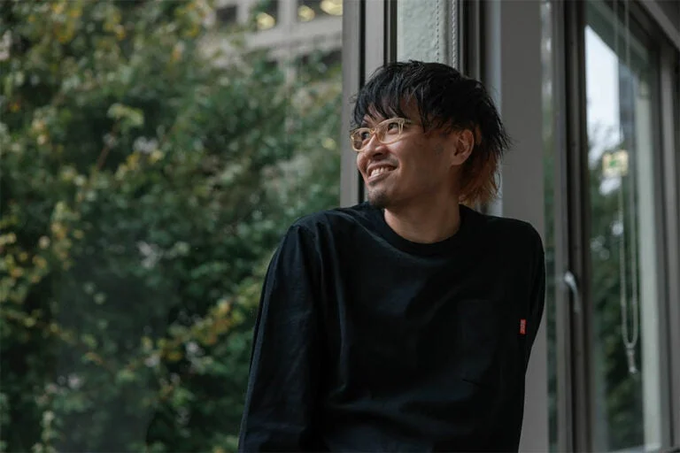 窓際に座り、笑顔で外を見つめる男性のポートレート。黒い長袖シャツとメガネが特徴的で、背景には緑豊かな木々が見える。リラックスした雰囲気です。