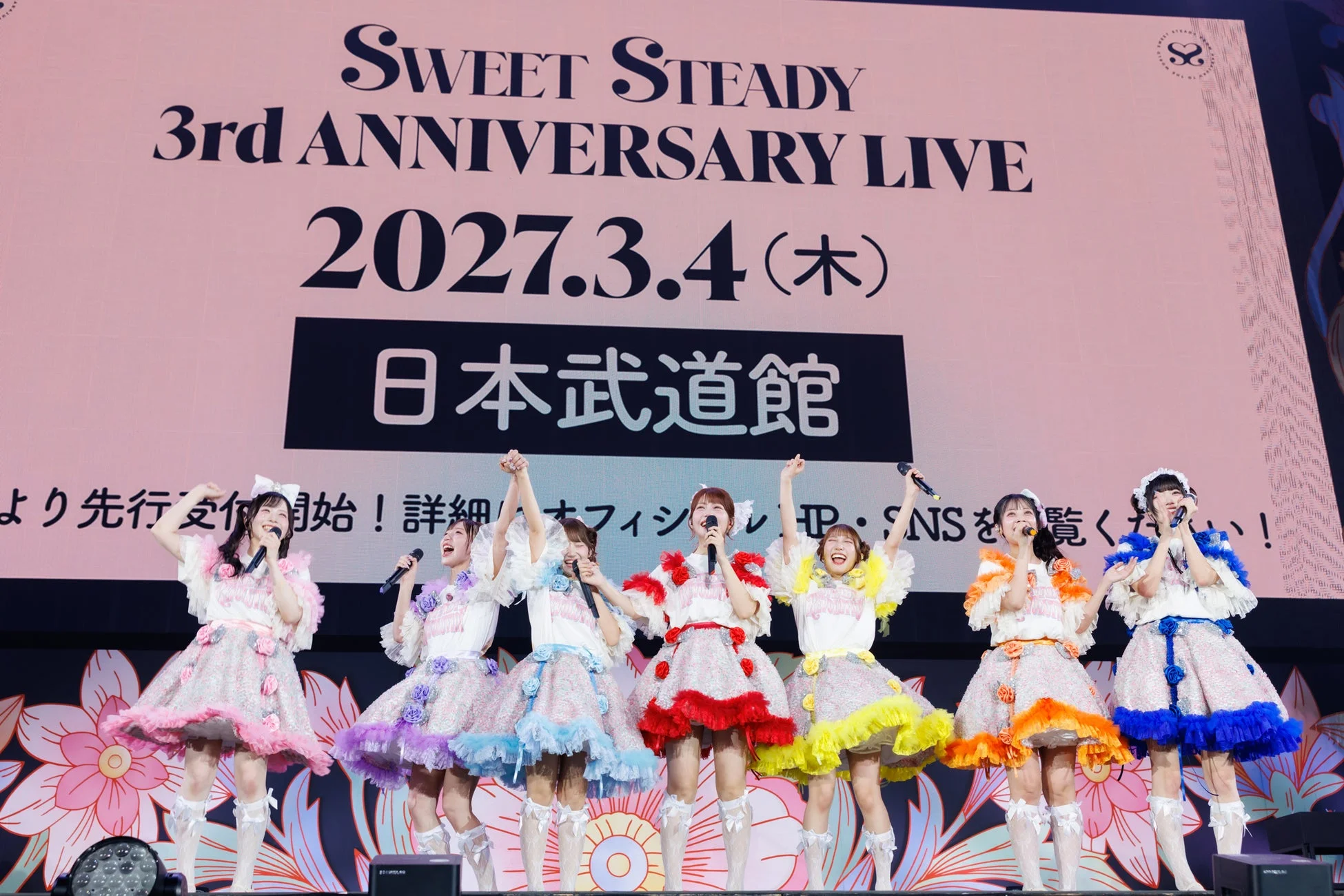 アイドルグループSweet Steadyの3周年記念ライブの様子。カラフルなフリル衣装をまとったメンバー7人がステージで歌唱しており、背景スクリーンには「SWEET STEADY 3rd ANNIVERSARY LIVE 2027.3.4 (木) 日本武道館」と表示されている。