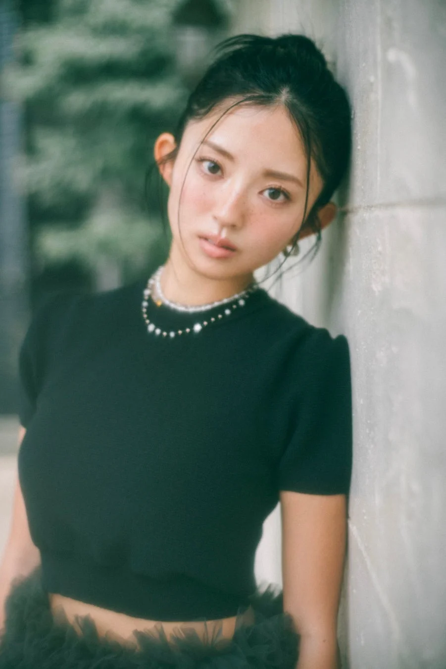黒いトップスとスカートを着用し、パールネックレスをつけた若い女性のポートレート。壁に寄りかかり、カメラを見つめている。そばかすが特徴的で、背景は緑がかったぼかしとなっている。