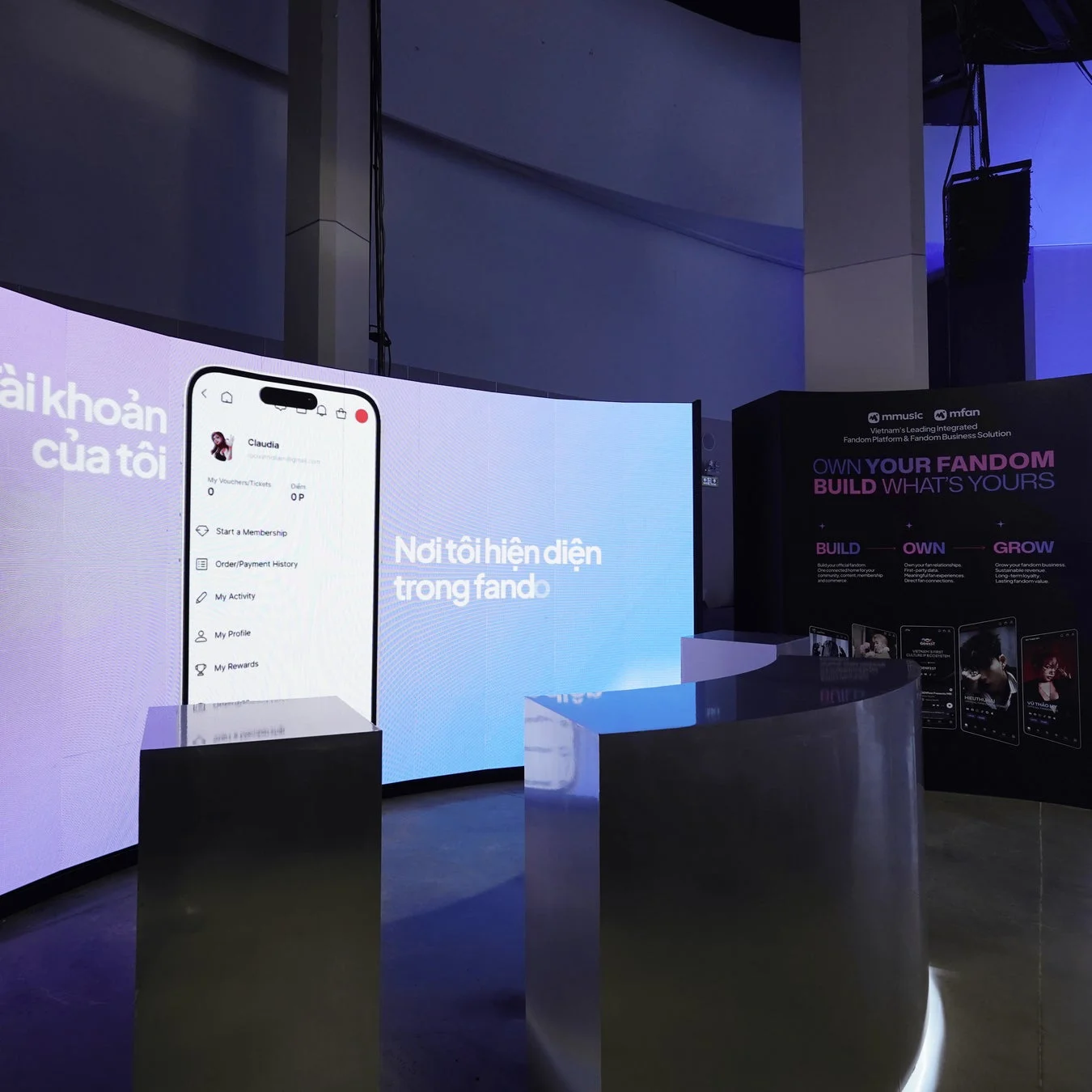 ファンダムプラットフォームの展示ブースの様子を捉えた画像です。湾曲したLEDスクリーンにモバイルアプリのインターフェースやベトナム語のテキストが表示され、右側のバナーには「OWN YOUR FANDOM BUILD WHAT'S YOURS」というメッセージとサービスの説明が書かれています。