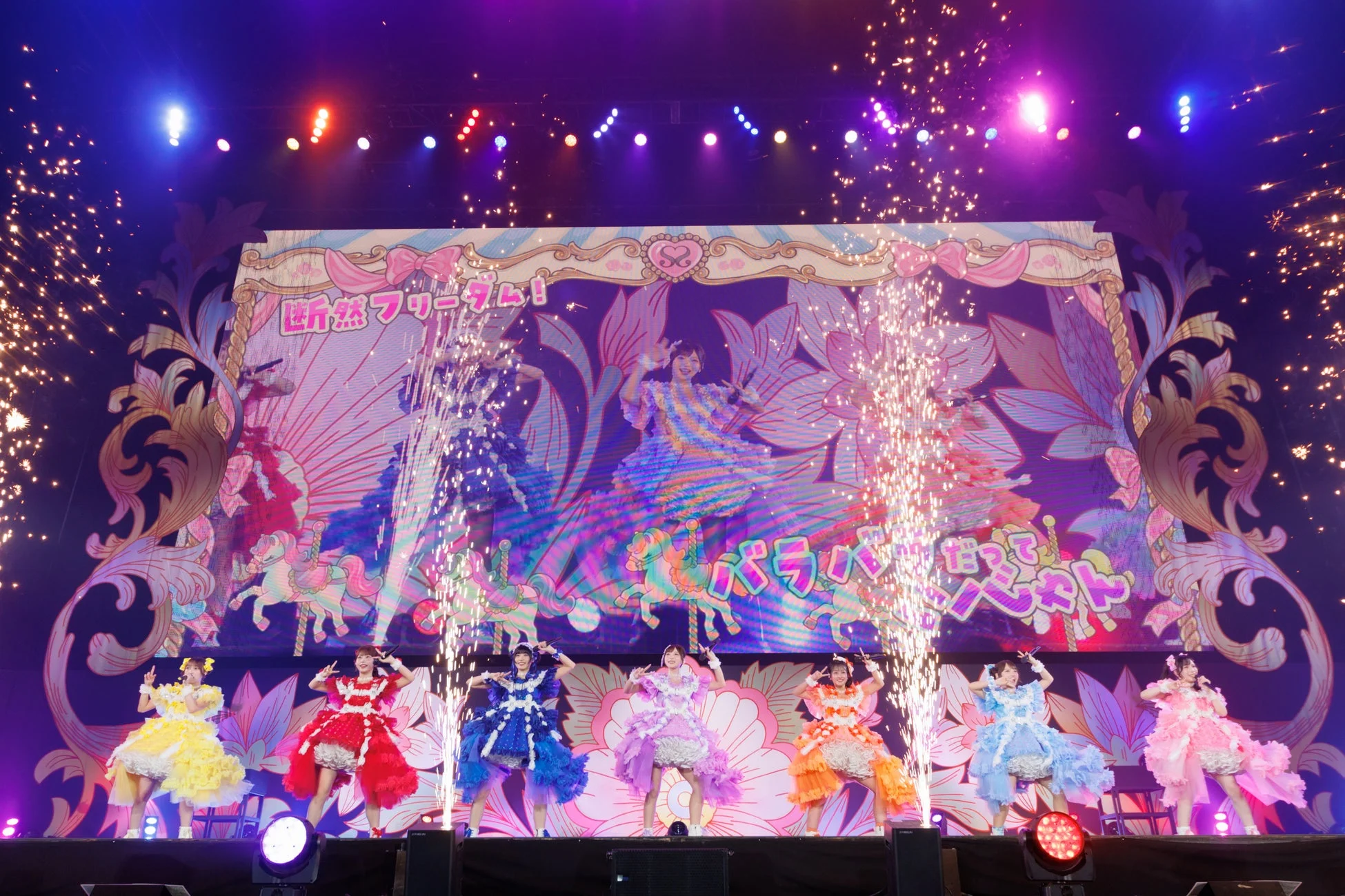 カラフルな衣装をまとったアイドルグループがステージで歌い踊るライブパフォーマンス。背景には花やメリーゴーランドが描かれたスクリーンがあり、火花が噴き出す華やかな演出で熱気に包まれている。