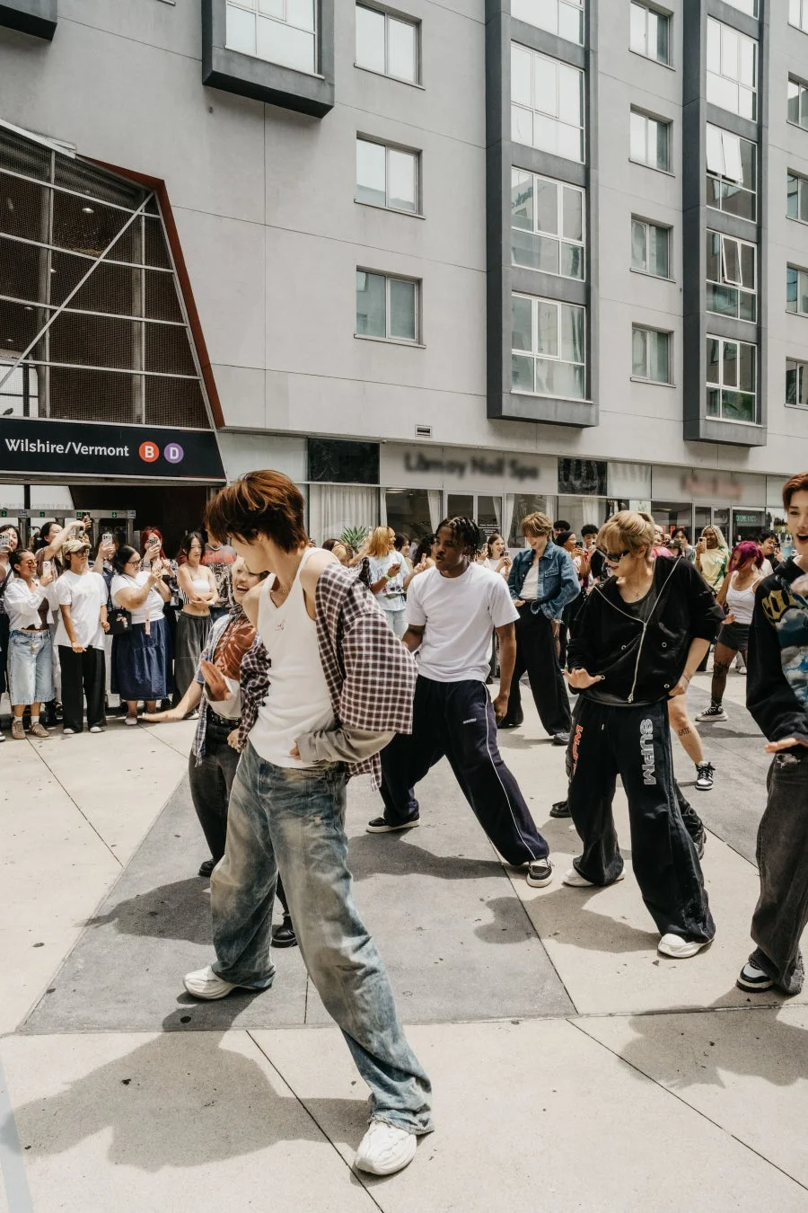都市の通りで若者たちがストリートダンスパフォーマンスを披露しており、多くの観客がそれを見守っています。背景には地下鉄の駅入り口や建物が見え、活気あるイベントの様子がうかがえます。