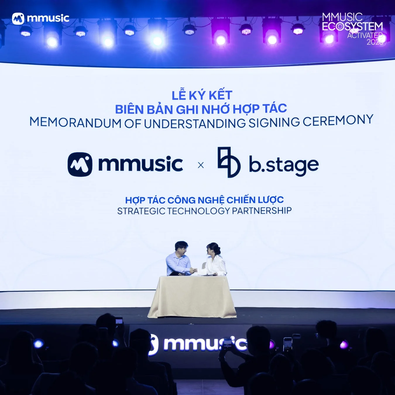 mmusicとb.stageによる戦略的技術提携の覚書調印式が開催されている様子です。ステージ上では二人の代表者が着席しており、聴衆が見守る中で契約が交わされています。背景にはイベントタイトルが表示されています。