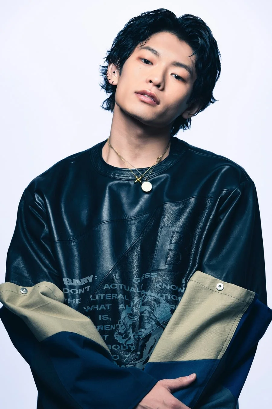 白い背景のスタジオで撮影された、黒いレザートップスを着用した若い男性のポートレートです。複数のネックレスを身につけ、クールな表情でこちらを見ています。