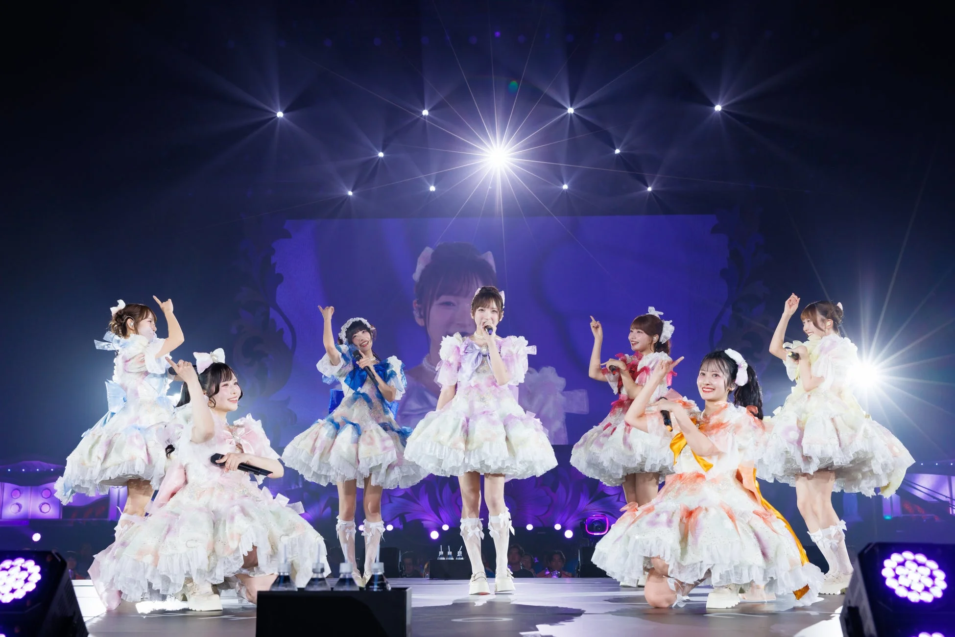 明るいステージ照明の下、カラフルでフリルの多い衣装を身につけた複数の女性アイドルがパフォーマンスを行っています。マイクを手に歌い、笑顔で観客を魅了している様子がうかがえます。背景にはメンバーの顔が映し出された大きなスクリーンがあります。