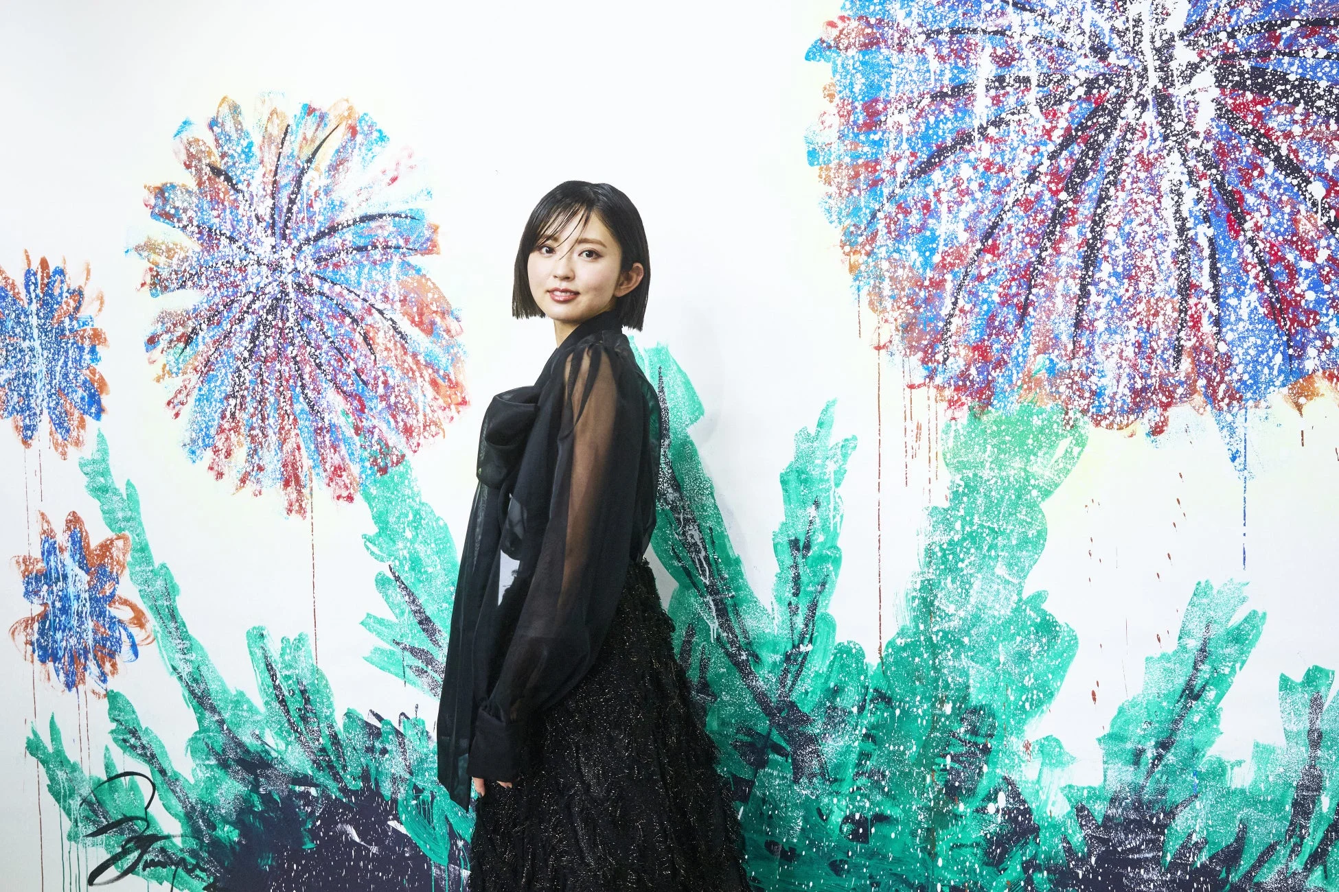 黒いシアーな衣装をまとった短髪の女性が、カラフルな抽象画が描かれた白い壁を背景に優しく微笑んでいるポートレート写真です。