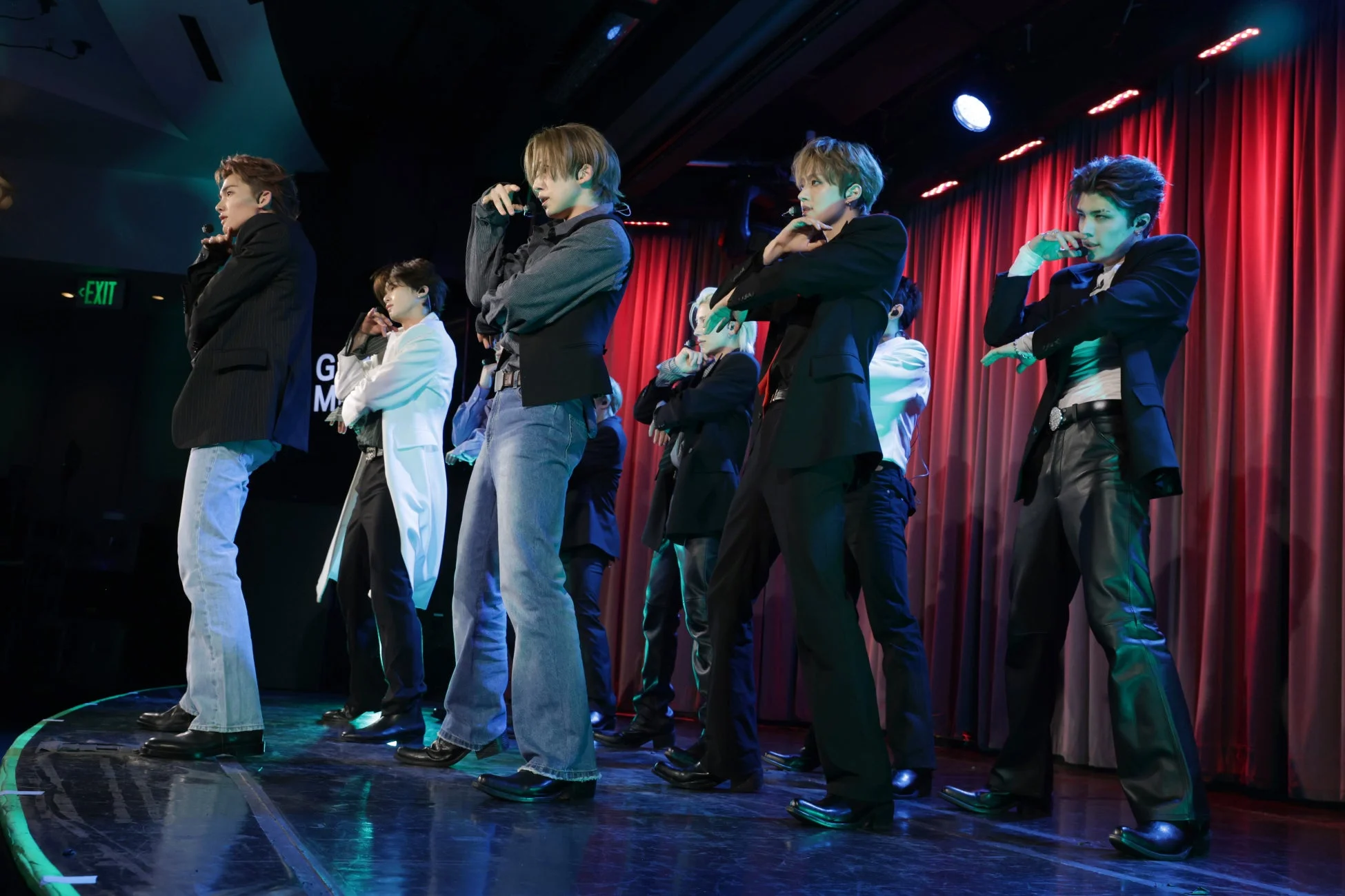 男性アイドルグループがステージ上でパフォーマンスを行っている瞬間を捉えた画像です。メンバーは様々なスタイルの衣装を身につけ、ポーズをとっており、背景には赤いカーテンが見えます。