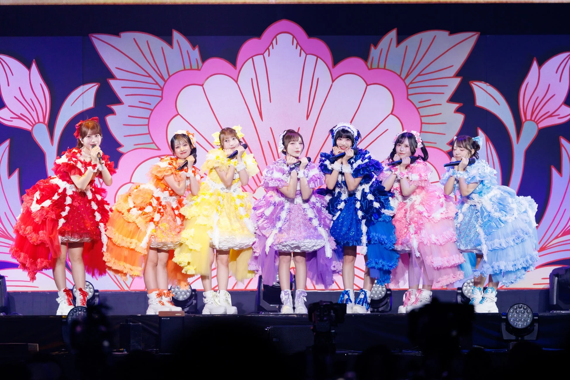 華やかなフリルドレスを着用した7人の女性アイドルグループが、マイクを手にステージでパフォーマンスしている様子です。背景には大きな花柄の装飾があり、明るく楽しいライブの雰囲気が伝わってきます。