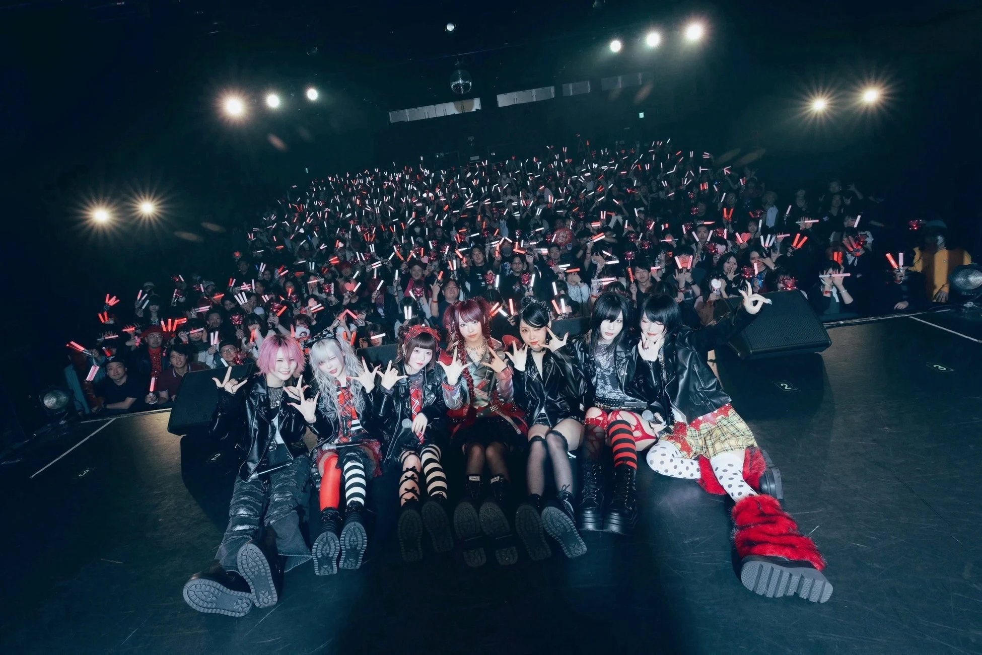 ライブ会場のステージで、個性的なファッションの7人組パフォーマーが、無数のペンライトを掲げる観客を背景にポーズ。