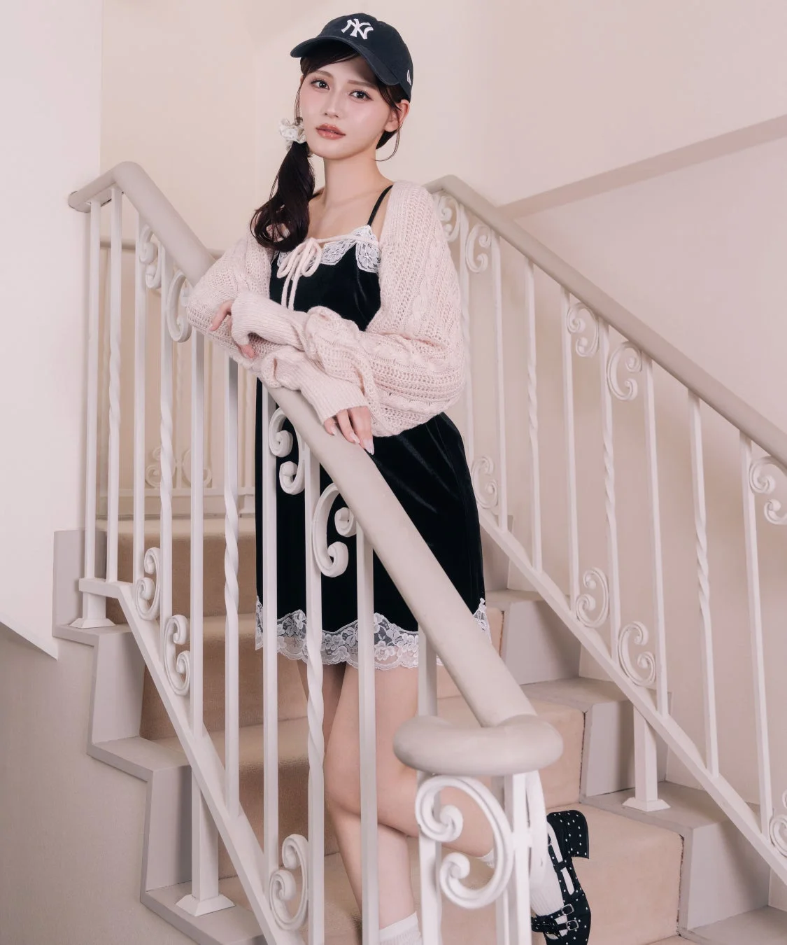 黒いベロアのキャミワンピースとピンクのニットボレロを着た女性が、NYロゴのキャップを被り、白い手すりの階段に立っています。可愛らしいカジュアルなファッションです。