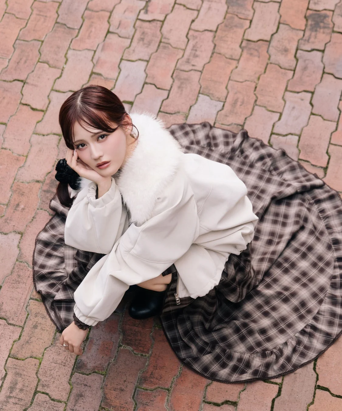レンガ調の地面に座り込み、カメラを見つめる若い女性のポートレート。白いファー付きの襟のコートと茶色のチェック柄のスカートを着用し、頬に手を添えたポーズをとっています。
