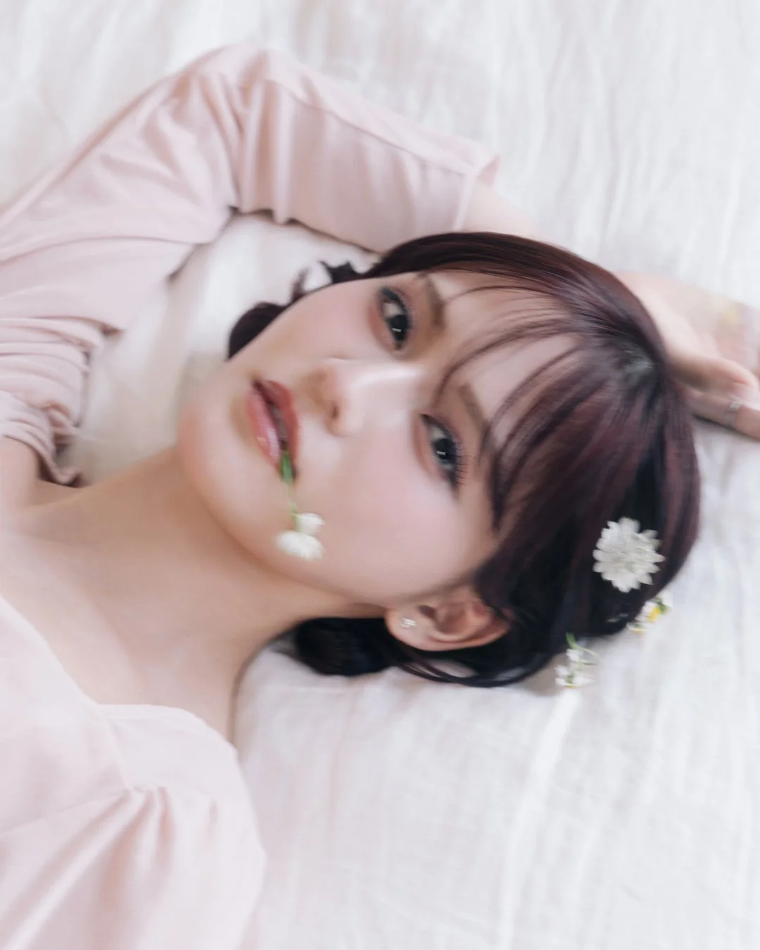 ピンク色の服を着た女性が白いシーツに横たわり、髪や口元に小さな白い花を飾っているポートレートです。全体的に柔らかく、幻想的な雰囲気が漂っています。