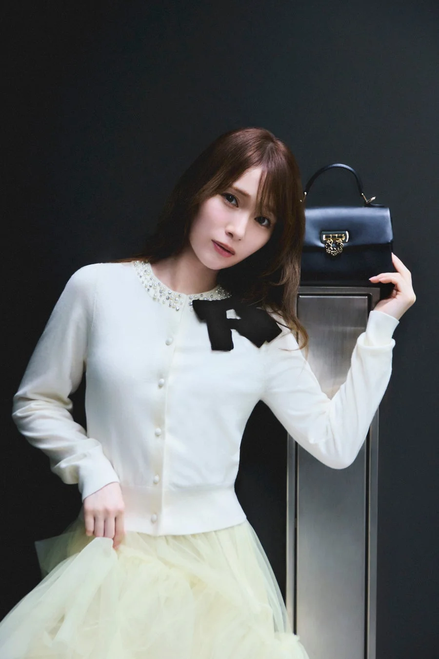 白いカーディガンと淡い黄色のチュールスカートを着用した女性が、黒いハンドバッグを手にポーズ