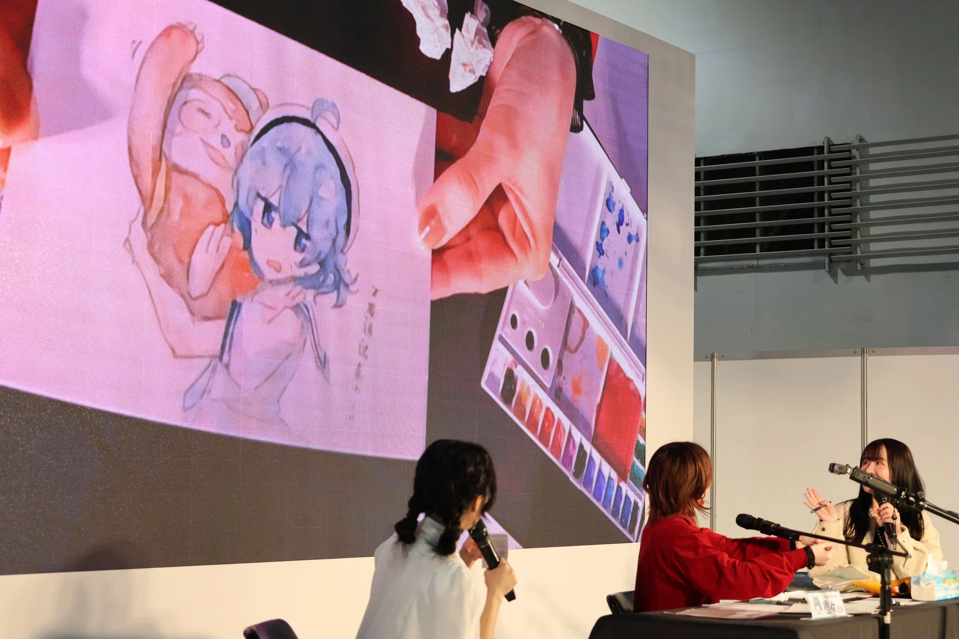 イベント会場で、3人の登壇者がマイクを前にトークセッションを行っている。スクリーンには水彩画のアニメキャラクターと水彩パレットの映像が映し出されており、アート関連のプレゼンテーションやワークショップのようだ。
